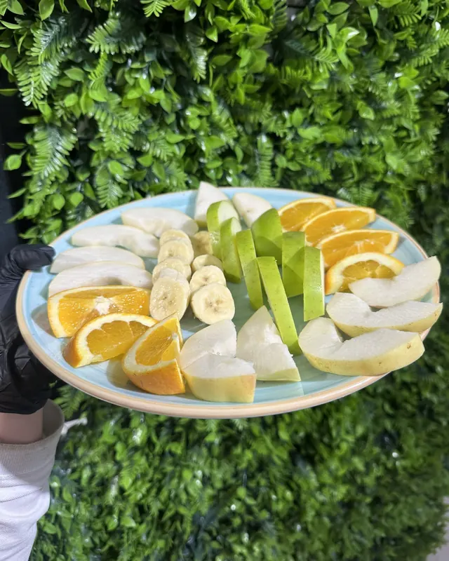 A plate of sliced fruit, orange, apple, banana and pear, held up against a green wall